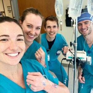 Dr. Kimberly Veltman and 3 classmates smile for a picture together in a clinical setting. Two of them hold up their wrists to show off their intravenous lines.